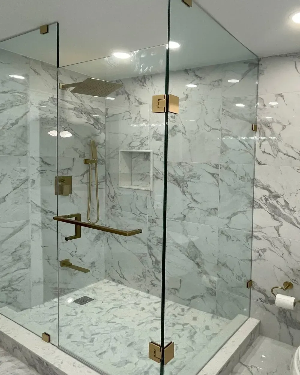 Close-up of a frameless glass shower door with brushed brass hinges and a brass towel-bar handle against marble-look tile